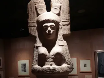 The Young Woman of Amajac sculpture displayed on a white pedestal in a dim museum hall.