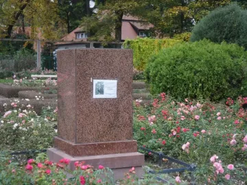 Brown granite base of The Dancing Girl monument with a small placard, surrounded by flowering bushes and trees.