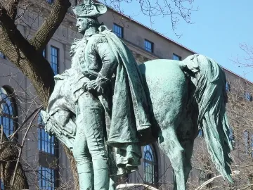 Verdigris-patinated George Washington sculpture beside a horse, seen from a low angle with bare branches and a building behind it.
