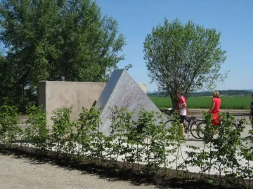 Hailfingen-Tailfingen Memorial beside a concrete wall, with two people and bicycles nearby, shrubs lining a gravel path, and fields and trees in the background.