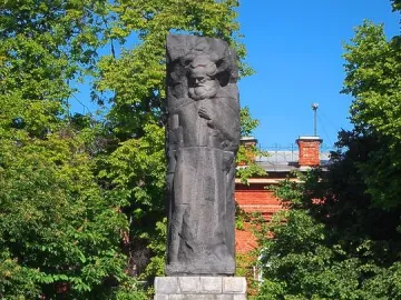 Karl Marx monument with a dark stone figure on a tiered base, framed by green trees and a brick building.