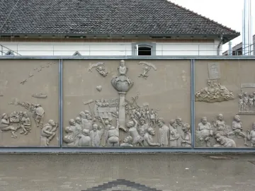 Ludwigs Erbe stone relief with three panels showing a crowded central monument scene and smaller figures on either side.