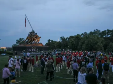 The Marine Corps War Memorial stands illuminated at dusk with bronze figures raising a flag, while a crowd gathers on the lawn below.
