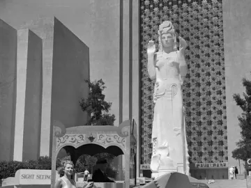 Pacifica statue of a woman with raised hands and an elaborate headdress stands before a star-patterned wall, with two women posing beside a sightseeing vehicle.