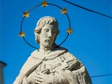 Sandstone statue of St. John of Nepomuk with a halo of stars against a blue sky.