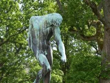 The Shade bronze sculpture of a nude male figure leaning forward on a rock in a garden setting.