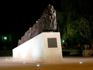 The Train of Pain monument at night, showing a long dark sculpture of clustered human figures on a light-colored base.