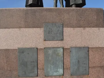 Bronze name plaques on the granite base of the Auswandererdenkmal, with the lower legs of the figures visible above.
