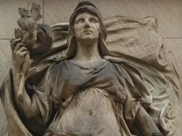 Weathered stone relief on the Commodore John Barry Statue showing a draped female figure with a head covering, holding a leafy branch and resting a hand on a sword hilt.