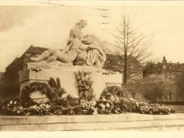 Stone war memorial with two sculpted figures embracing on a pedestal, surrounded by wreaths and flowers.