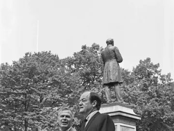Two men in suits walk in front of the Johan Ludvig Runeberg statue in a park.