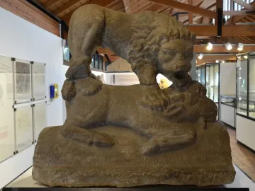 The Corbridge Lion sandstone sculpture showing a lion standing over another animal, displayed indoors beside other stone objects.