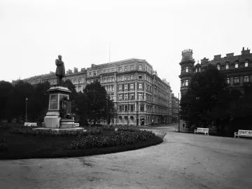 Johan Ludvig Runeberg statue on a large stone pedestal along a park pathway, with historic buildings and trees behind it.