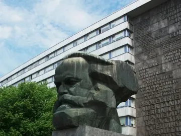 Karl Marx Monument, a large dark bust on a tall granite pedestal, with a modern building behind it.