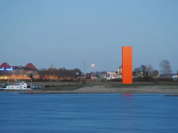 Rhine Orange sculpture standing on the far bank of a wide river with a distant townscape behind it.