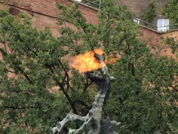 Wawel Dragon sculpture spewing a bright flame from its open mouth beside trees and a brick building