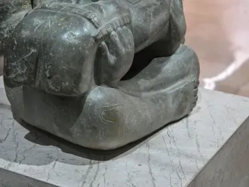 Lower body of the Las Limas Figurine seated on a marble base, with bent legs, visible feet, and scratch marks on the stone surface.