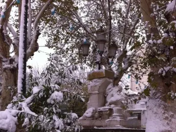Ain El Fouara Fountain covered in snow, with sculpted figures, a street lamp on top, and snow-laden trees and fence nearby.