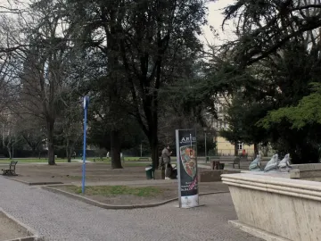 Stone fountain with metal frog statues beside a park path, with trees and a building in the background.