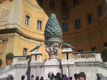 La Pigna bronze pine cone sculpture on a carved pedestal with peacock statues in front of a curved building.