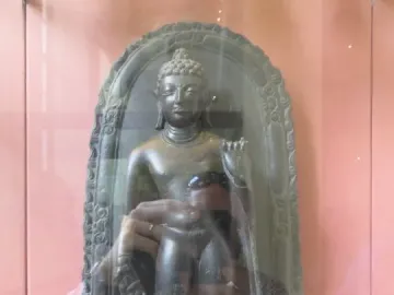 Sultanganj Buddha displayed in a glass case, with a standing figure inside an arched stele and a kneeling figure at the base.