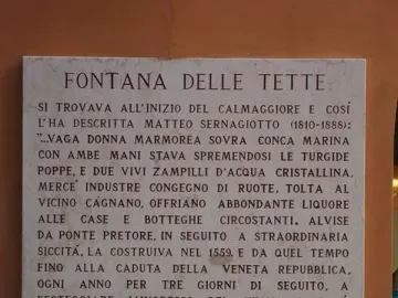 Marble plaques on a reddish-brown wall beside the Fontana delle Tette, with one plaque in uppercase text and a smaller cursive plaque below.