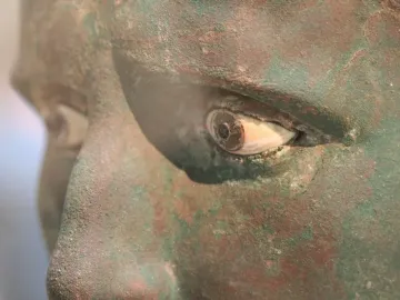 Close-up of the Meroë Head showing the eye, nose, and cheek of a weathered bronze statue with green and reddish-brown patina.