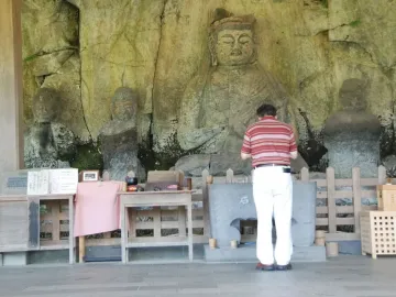 A man bows before the Usuki Stone Buddhas carved into a rock wall, with offerings placed on tables in front of the figures.