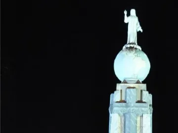 Divino Salvador del Mundo monument illuminated at night, with a robed statue standing on a glowing blue globe above a tall stone pedestal.