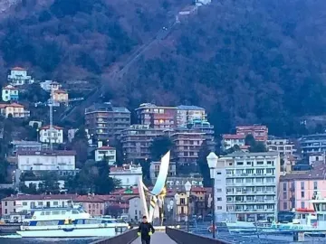 Life Electric sculpture standing at the end of a pier with a lakeside town and mountains behind it.