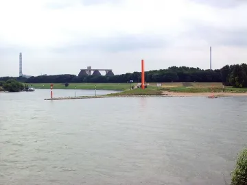Rhine Orange sculpture standing on a grassy riverbank beside a wide river, with trees and industrial structures in the background.
