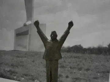 Small bronze model of the Monument to Stjepan Filipović with raised clenched fists on a pedestal, shown before a large black-and-white background photo of the monument.