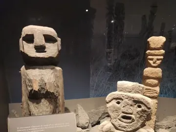 Chemamull wooden figure displayed among grey stones with smaller stone sculptures nearby.