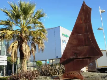 Dorna rust-colored metal sculpture with a sail-like curved shape in a landscaped area with palm trees and a white building behind it.