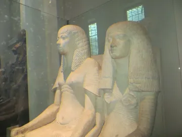 Horemheb and Amenia seated side by side in a light-colored ancient Egyptian sculpture behind glass.
