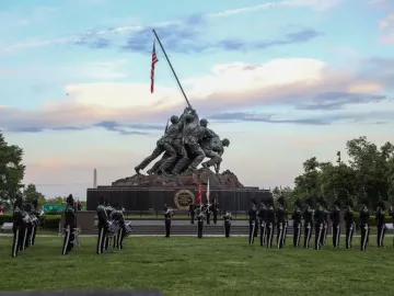 The Marine Corps War Memorial at twilight, with a military band or honor guard assembled on the lawn in front of the statue.