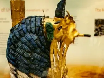 Ram in a Thicket sculpture with dark curved horns, blue and white body inlay, and a gold-colored support in a museum display.