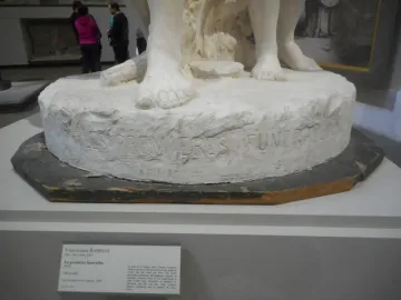 Close-up of The First Funeral sculpture showing the carved inscription and several bare feet on the plaster base.