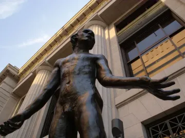 Bronze statue of Jean de Fiennes with arms outstretched, seen from below against a stone building.