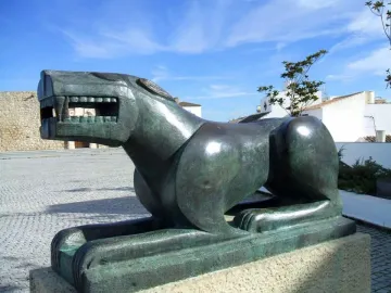 The Leona de Baena sculpture in green metal, shown in profile on a stone base in a town square.