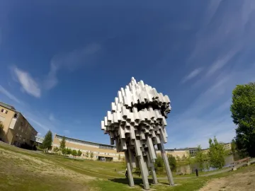 Norra skenet sculpture with clustered silver blocks beside a grassy lawn, with buildings, water, and trees in the background.