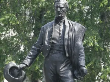 Bronze statue of General José Gervasio Artigas standing in historical clothing and holding a hat, with trees and a building behind it.