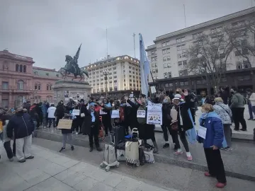 Manuel Belgrano equestrian monument on a tall stone pedestal behind a crowd in Plaza de Mayo, with Argentine flags and the pink Casa Rosada in the background.