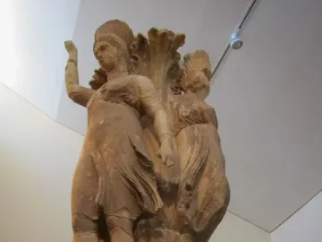Dancers of Delphi sculpture showing two draped female figures with raised arms around a central pillar on an ornate base.