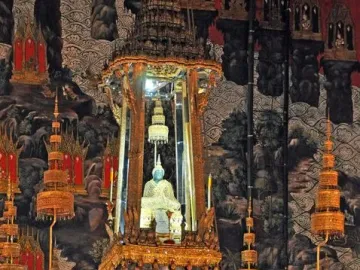 Emerald Buddha statue in a pale golden robe on an ornate golden altar, surrounded by gilded decorations and mural-covered walls.