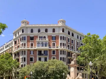 The Fountain of Hercules stands on a tiered stone base in a Barcelona square with trees and a large building behind it.