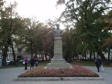 Bust of Pushkin on a tall stone pedestal in a park with pink flowers, trees, and people walking nearby.