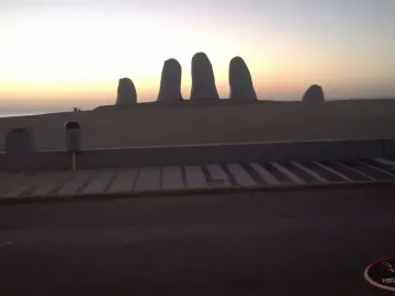 The Hand sculpture rising from a sandy beach at sunset, with the ocean in the background.