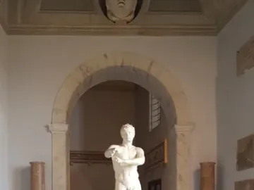 Apoxyomenos, a white marble nude male statue, stands on a pedestal in a museum room with an arched entryway and tiled floor.