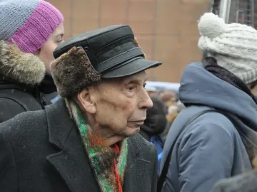 An elderly person in a fur-lined cap and striped scarf stands near the Solovetsky Stone in snowy weather.